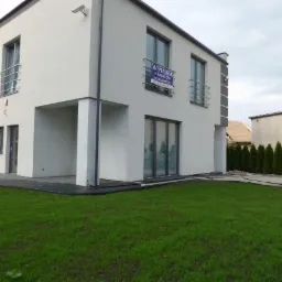 Nowoczesny, dwukondygnacyjny dom z białym tynkiem, szarymi detalami i balkonami z metalowymi balustradami, z tabliczką informacyjną o wynajmie na elewacji, otoczony trawnikiem i tujami.