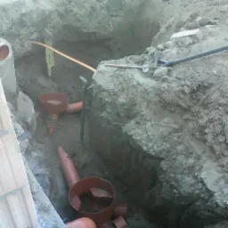 Wykop z widocznymi rurami kanalizacyjnymi w trakcie instalacji, częściowo zasypany ziemią, z narzędziami na górze wykopu. Ceglana ściana obok.
