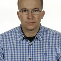 Portret mężczyzny w okularach i niebieskiej koszuli w kratę z haftowanym logo na piersi.