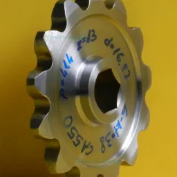 Metalowe koło łańcuchowe z niebieskimi oznaczeniami technicznymi na ż&oacute;łtym tle. Widoczne detale obr&oacute;bki i specyfikacja produktu.