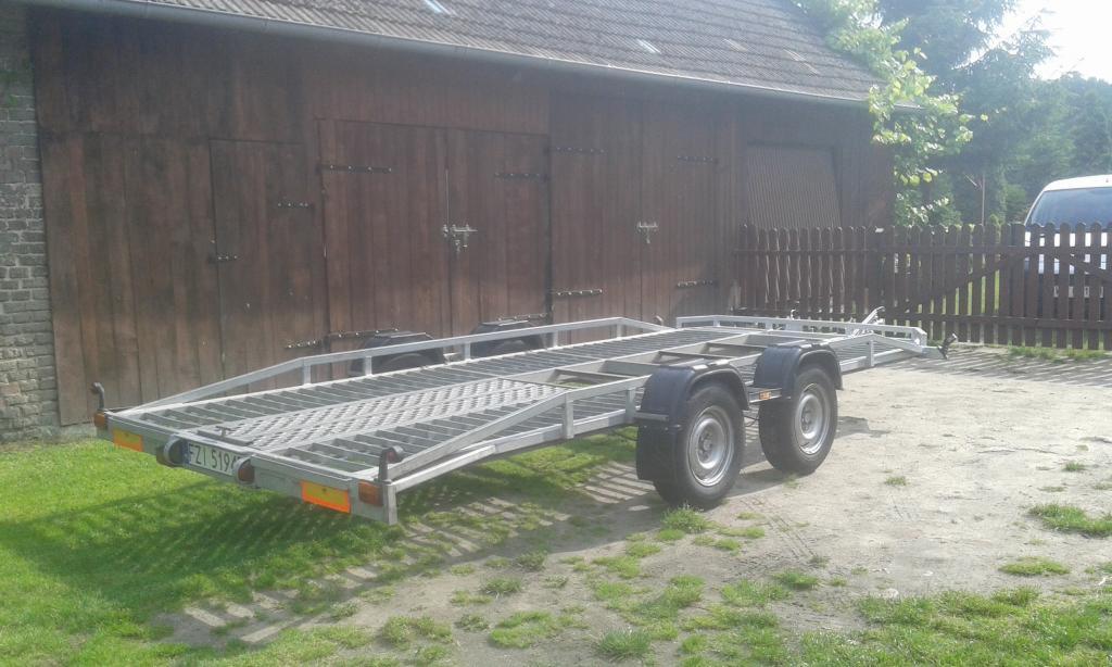 Pusta, dwuosiowa laweta z metalową kratownicą na platformie, zaparkowana na żwirowym podłożu przed drewnianym budynkiem gospodarczym i częściowo widocznym ogrodzeniem.