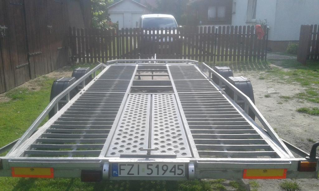 Pusta, aluminiowa laweta samochodowa z widoczną tablicą rejestracyjną FZI 51945, zaparkowana na trawie przed drewnianym płotem i białym budynkiem w tle.