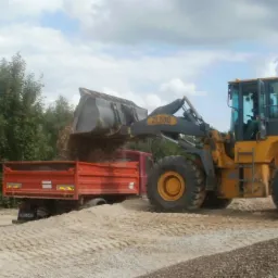 Żółta ładowarka ZL50G nasypuje materiał do czerwonej ciężarówki na tle drzew i pochmurnego nieba. Widoczne ślady opon na materiale.