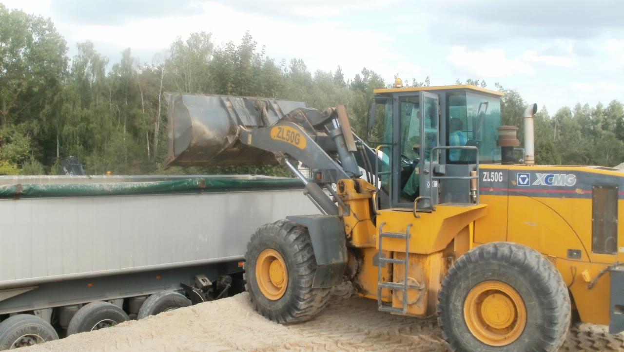 Żółta ładowarka XCMG ZL50G nabiera materiał do srebrnej naczepy, na tle drzew. Widoczne opony i kabina operatora. Materiał sypki.