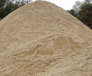 Góra jasnego piasku budowlanego z wyraźną fakturą, ułożona na zewnątrz, z drzewami w tle. Delikatne cienie podkreślają nierówności powierzchni.