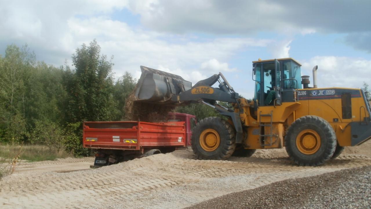 Żółta ładowarka ZL50G nasypuje materiał do czerwonej ciężarówki na tle drzew i pochmurnego nieba. Widoczne ślady opon na materiale.