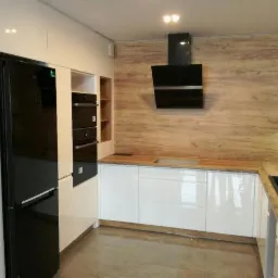 Nowoczesna kuchnia z białymi, lakierowanymi frontami szafek, drewnianym blatem i okładziną ścienną imitującą drewno; czarny okap i lodówka.