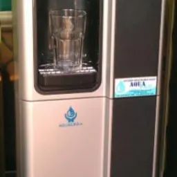 Dystrybutor wody z pustą szklanką, srebrno-czarna obudowa, naklejka '100' z czerwoną wstążką, butelka z napojem na górze.