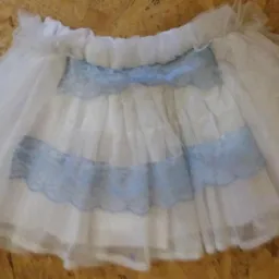 Biała, tiulowa spódniczka z falbanami i niebieskimi, koronkowymi wstawkami, leżąca na drewnianej podłodze.