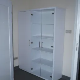 Biała szafa z przeszklonymi drzwiami i półkami w jasnym wnętrzu. Widoczne metalowe uchwyty i zawiasy. Obok drzwi wejściowe.