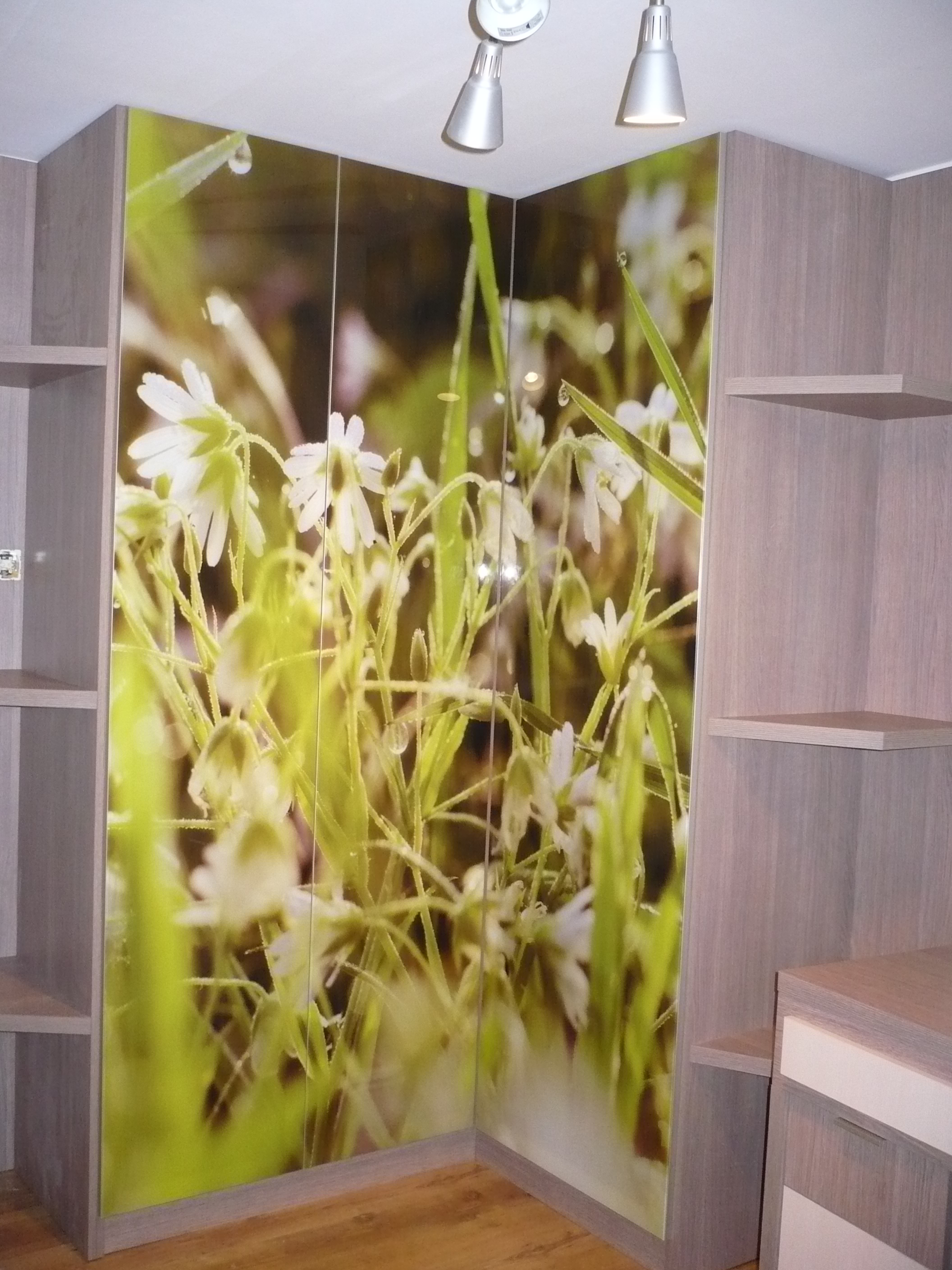 Szafa narożna z fototapetą przedstawiającą kwiaty i krople rosy, wkomponowana w jasne, drewniane meble z otwartymi półkami i oświetleniem punktowym.