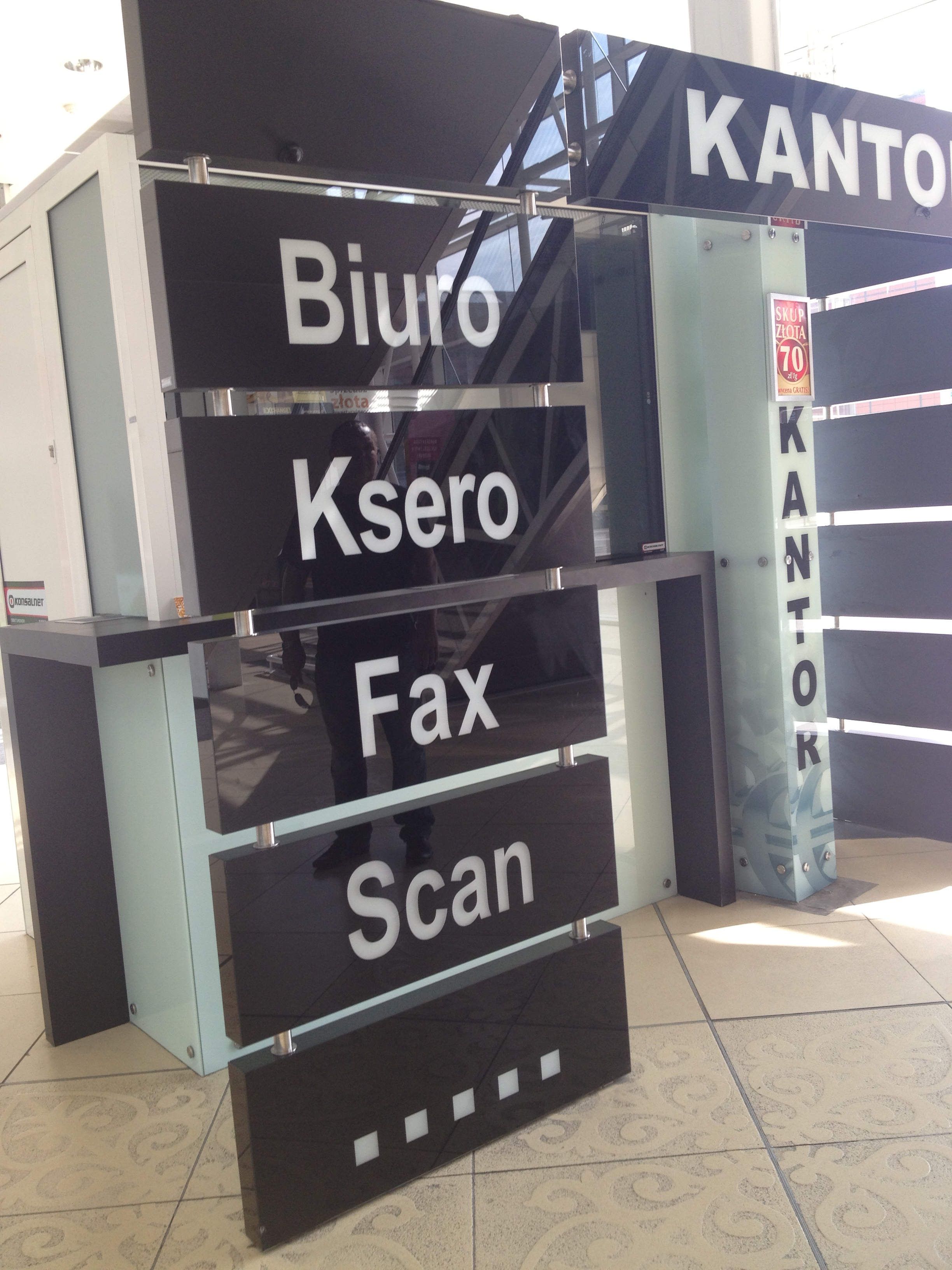 Nowoczesny, czarno-biały szyld z napisem 'KANTOR', 'Biuro', 'Ksero', 'Fax', 'Scan', wykonany ze szkła i drewna. Widoczna refleksja sylwetki osoby.
