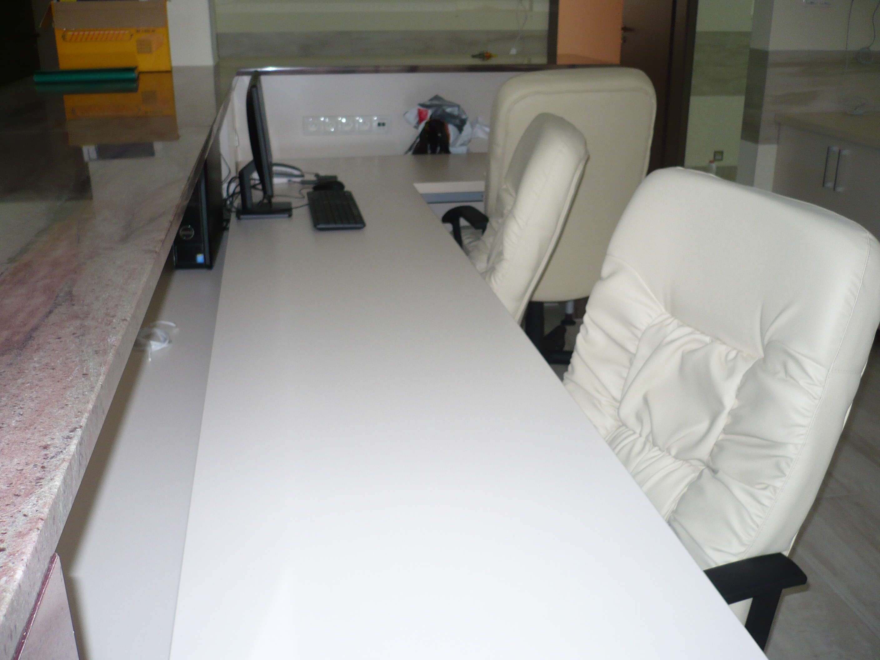 Długi, biały blat biurka z komputerem i dwoma białymi krzesłami w tle, oświetlony jasnym, naturalnym światłem. Blat wykończony marmurem.