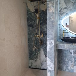 ROBBULID Robert Gawroński - Wykończona łazienka z kabiną prysznicową typu walk-in, wyłożona marmurowymi płytami w odcieniach beżu i zieleni, z złotą armaturą i podświetlanym lustrem.