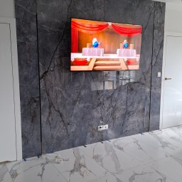 ROBBULID Robert Gawroński - Ściana z szarych płyt kamiennych z żyłami, na niej zawieszony telewizor. Podłoga wyłożona białymi płytkami z wzorem marmuru. Nowoczesne wykończenie wnętrza.