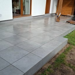 ROBBULID Robert Gawroński - Szara, kwadratowa, błyszcząca podłoga z płytek na zewnątrz budynku, z widocznym fragmentem trawnika i elementami drewnianej elewacji. Widok z niskiej perspektywy.