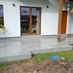 ROBBULID Robert Gawroński - Wykończony taras z szarych, kwadratowych płytek, z widocznym fragmentem ogrodu i elewacją domu z oknami. Część płytek jeszcze nieułożona.