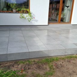 ROBBULID Robert Gawroński - Wyłożony szarymi płytkami taras z niskim stopniem, widok z dołu, zieleń w tle, okno z kwiatami na ścianie budynku. Minimalistyczny design.