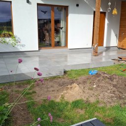 ROBBULID Robert Gawroński - Jasnoszara, kwadratowa płytka na tarasie domu z drewnianymi oknami i kolumną. W tle widoczny ogród z kwiatami i ziemią. Częściowo ukończona realizacja.