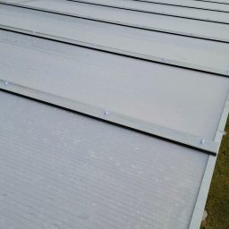 ROBBULID Robert Gawroński - Przezroczyste panele dachowe z widocznymi kroplami wody, zamontowane na metalowej konstrukcji, z fragmentem dachu budynku w tle.