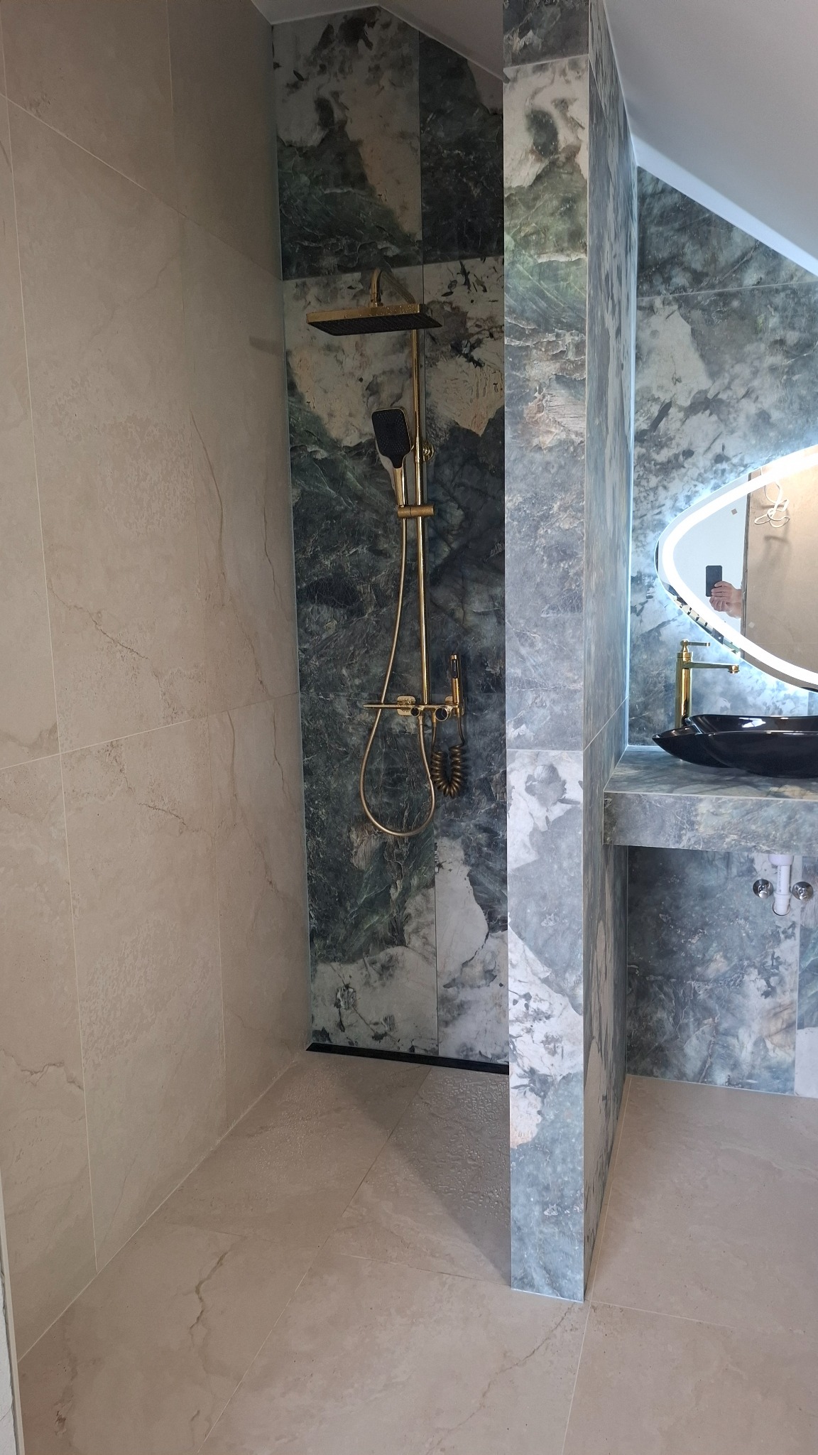 Wykończona łazienka z kabiną prysznicową typu walk-in, wyłożona marmurowymi płytami w odcieniach beżu i zieleni, z złotą armaturą i podświetlanym lustrem.