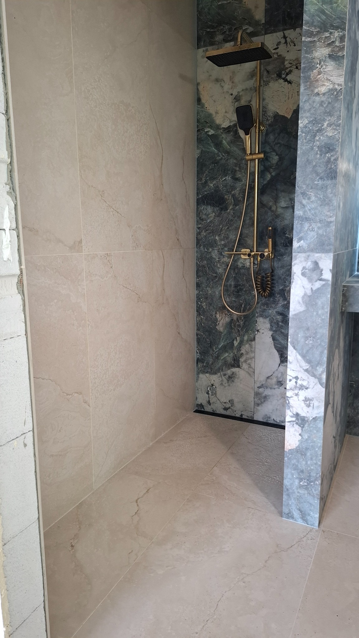 Wykończona kabina prysznicowa z beżowymi płytkami na ścianach i podłodze, oraz zielono-białymi płytkami imitującymi marmur pod złotą armaturą. Częściowo widoczny surowy mur.