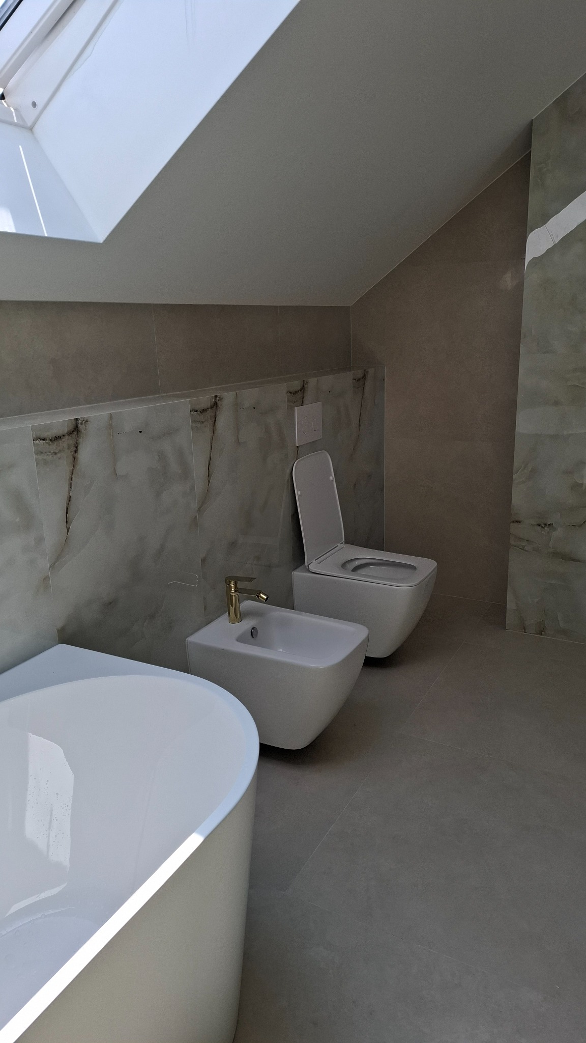 Wykończona łazienka na poddaszu: biała wanna, toaleta i bidet z złotą armaturą na tle marmurowych płytek i okna dachowego. Stonowana kolorystyka, nowoczesny design.