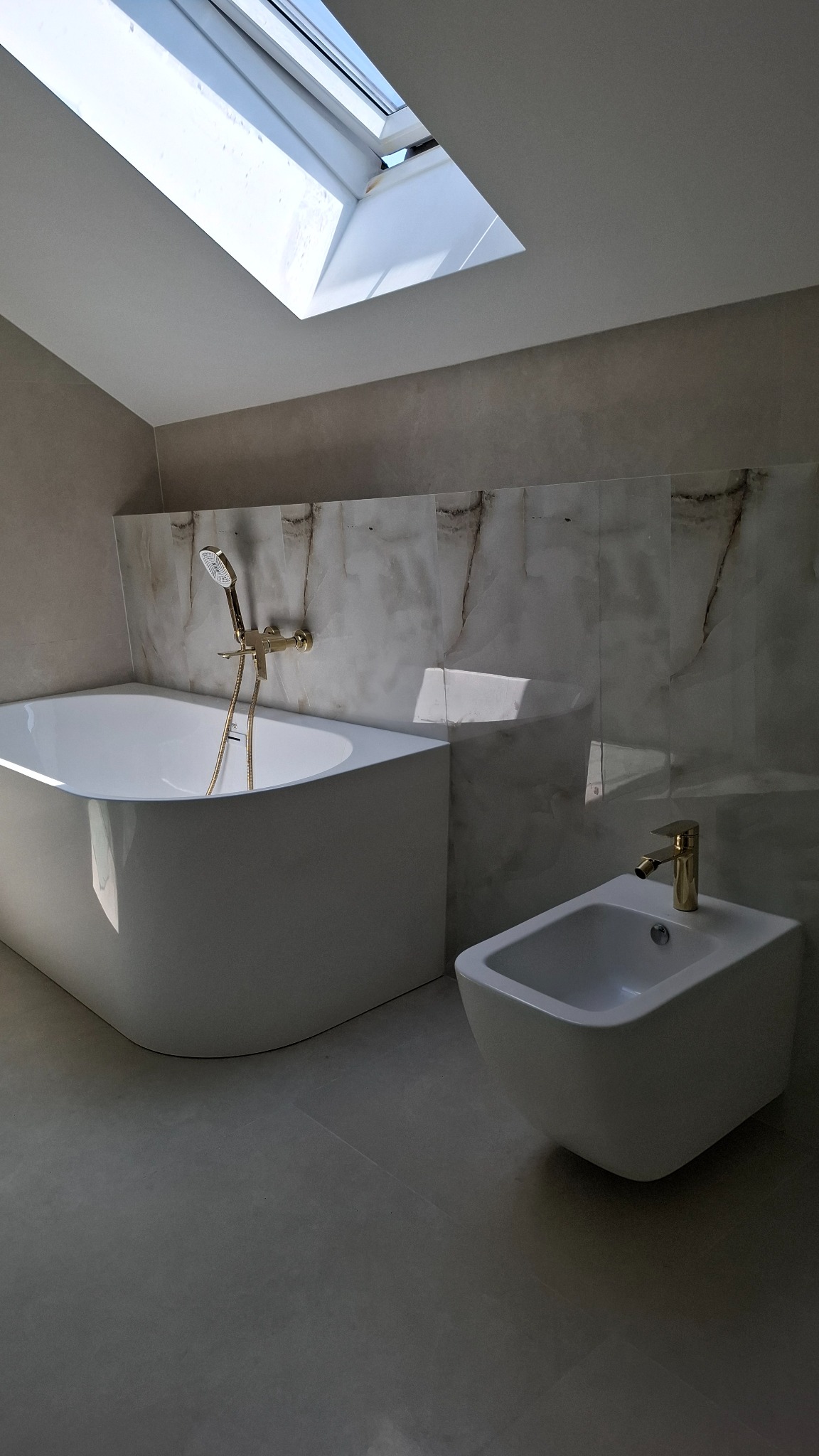 Minimalistyczna łazienka z wanną, bidetem i marmurowymi płytkami pod skosem sufitu z oknem dachowym. Armatura w kolorze złota.