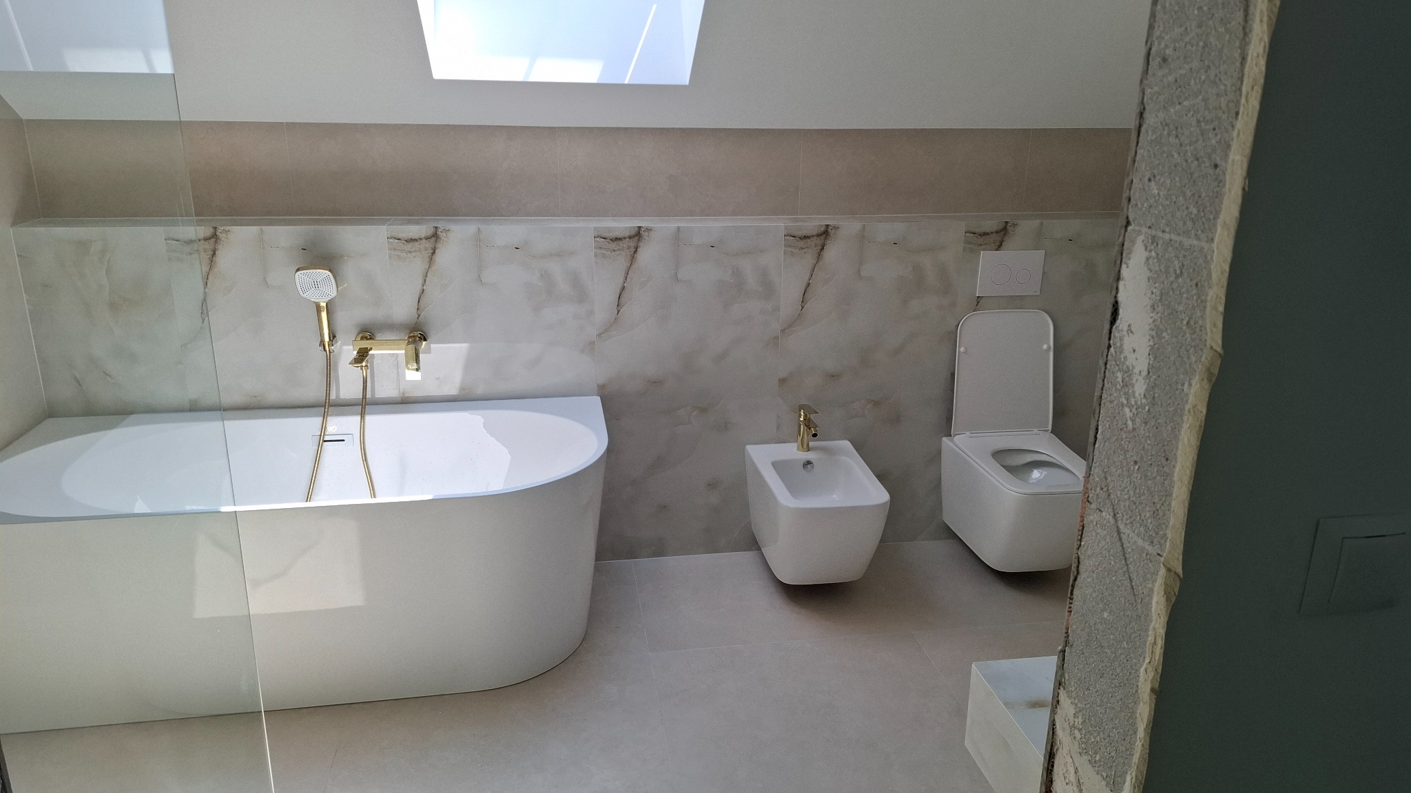 Wykończona łazienka z owalną wanną, bidetem i toaletą w stylu minimalistycznym. Beżowe płytki imitujące marmur na ścianach. Armatura w kolorze złota. Okno dachowe.