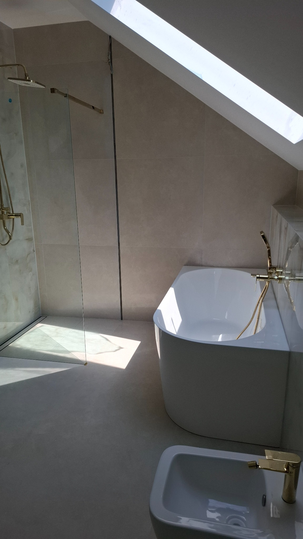 Jasna łazienka na poddaszu z kabiną prysznicową, wanną i bidetem. Złote baterie i detale. Okno dachowe wpuszcza naturalne światło.