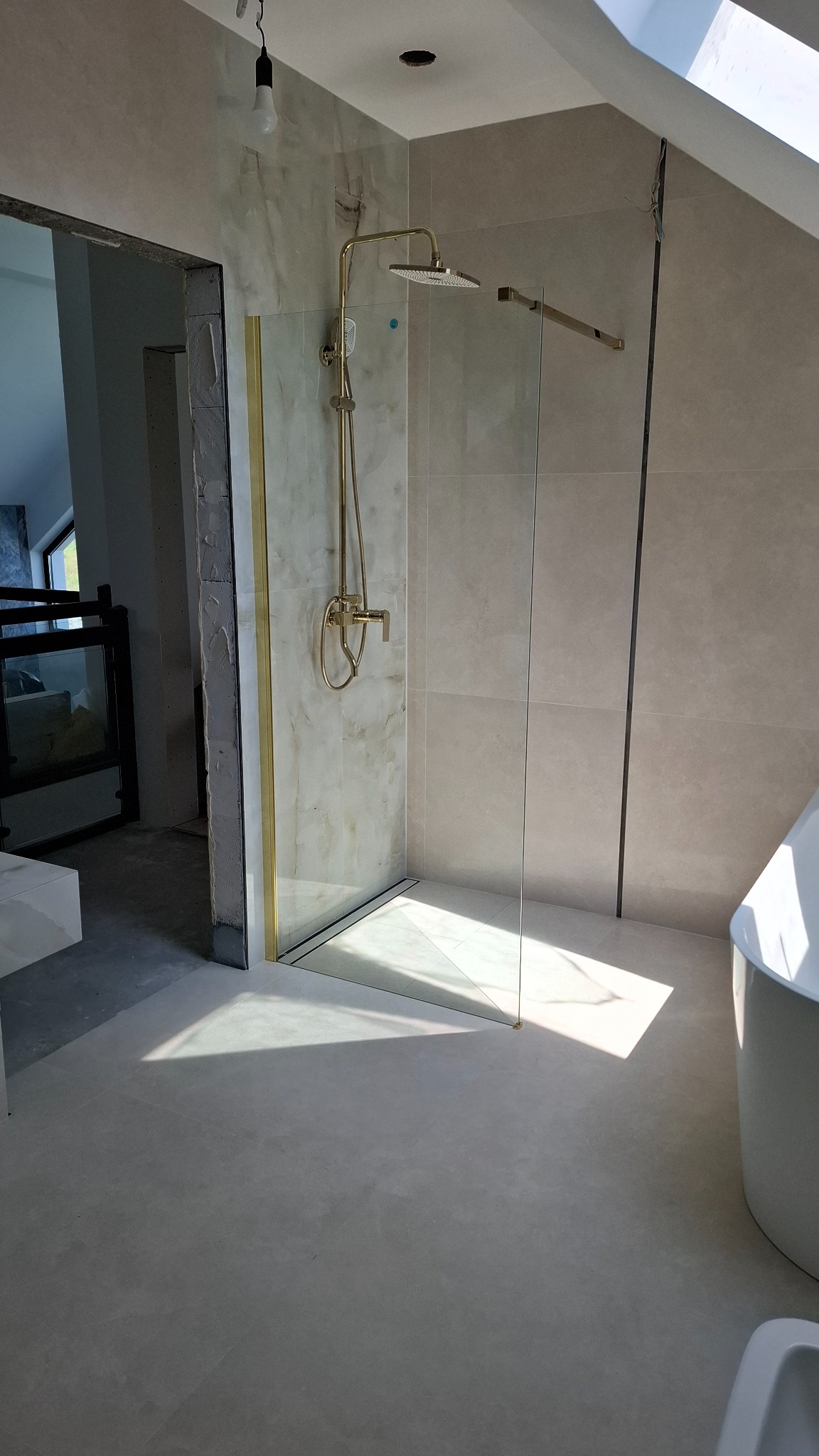 Beżowa łazienka w trakcie wykańczania: kabina prysznicowa ze złotymi detalami, widoczne płytki i fragment wanny. Otwarty kadr na sąsiednie pomieszczenie.