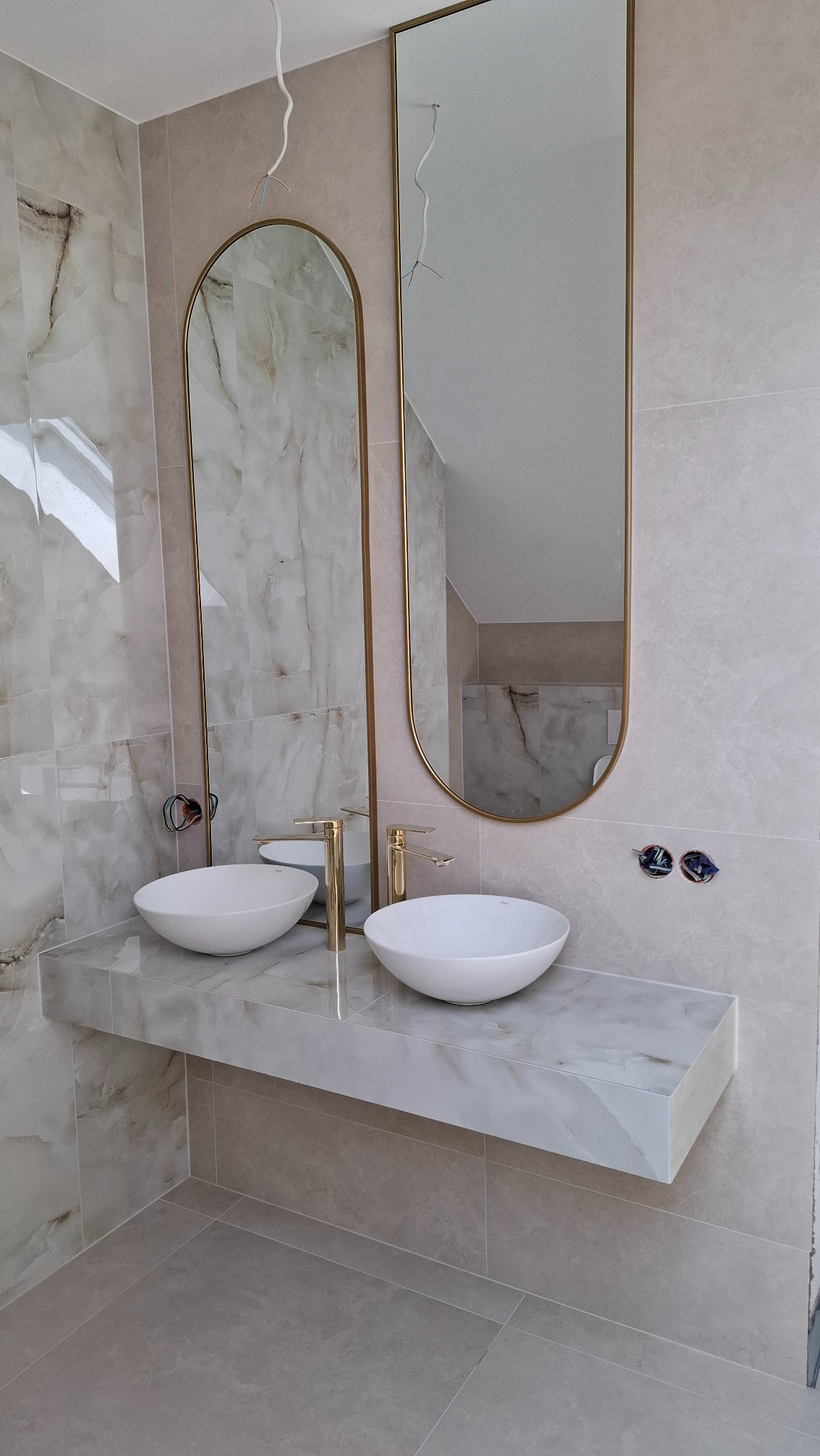 Wykończona łazienka z marmurowymi płytkami, dwiema owalnymi umywalkami i złotymi bateriami. Dwa wysokie lustra w złotych ramach. Widoczne kable od oświetlenia.