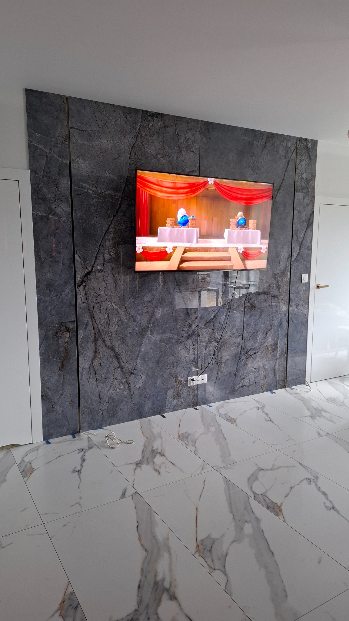 Ściana z szarych płyt kamiennych z żyłami, na niej zawieszony telewizor. Podłoga wyłożona białymi płytkami z wzorem marmuru. Nowoczesne wykończenie wnętrza.