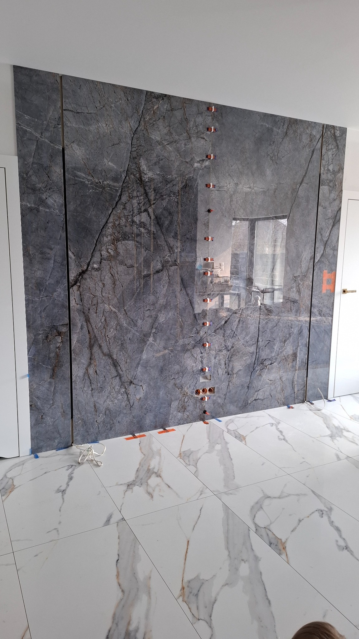 Montaż wielkoformatowych płytek imitujących marmur na ścianie, z użyciem poziomowania. Widoczne gniazdka elektryczne. Podłoga wyłożona jasnymi płytkami.