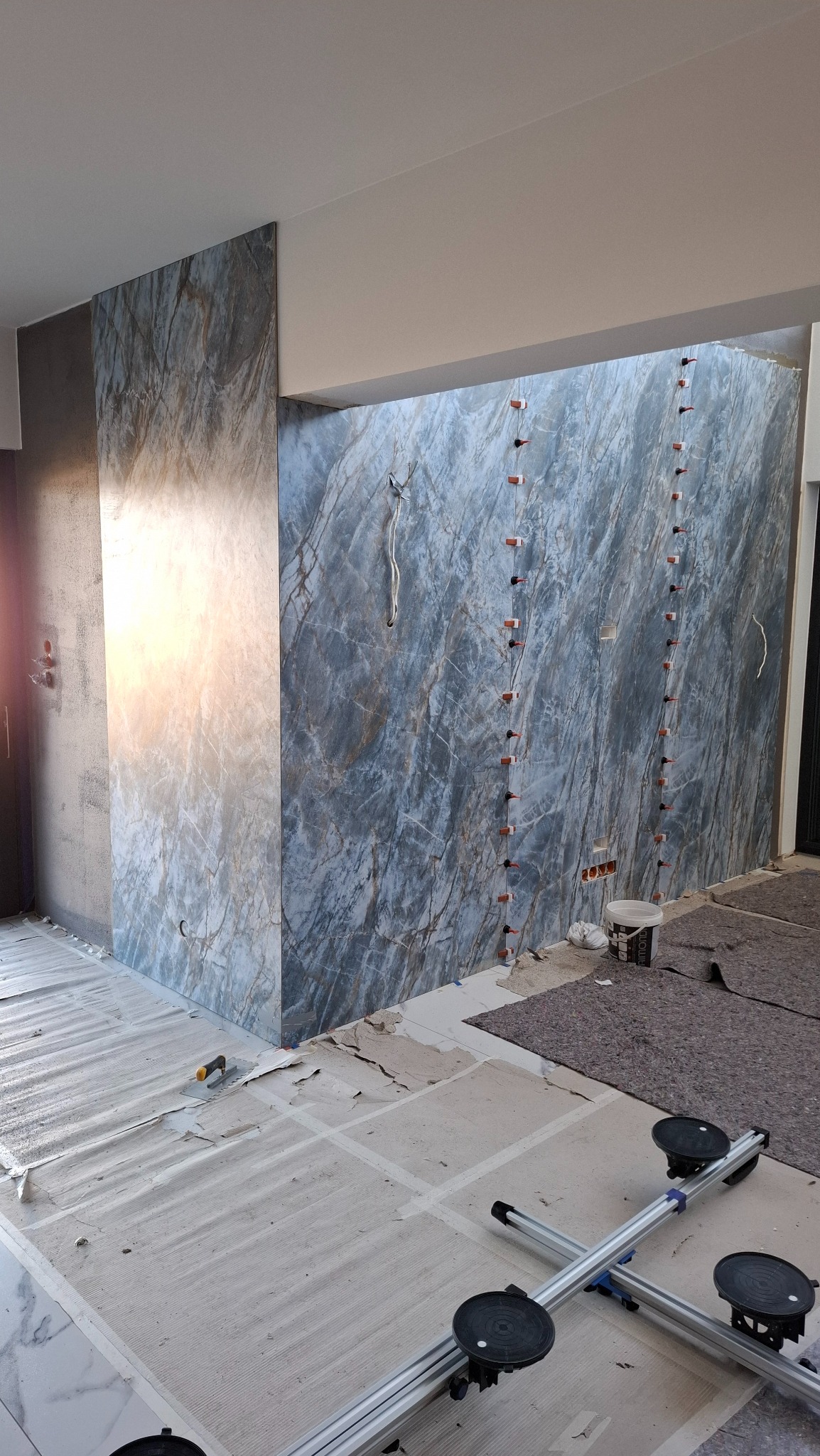 Montaż wielkoformatowych płytek imitujących marmur na ścianie, z użyciem systemu poziomowania. Widoczne narzędzia i fragment podłogi z zaznaczonym układem płytek.