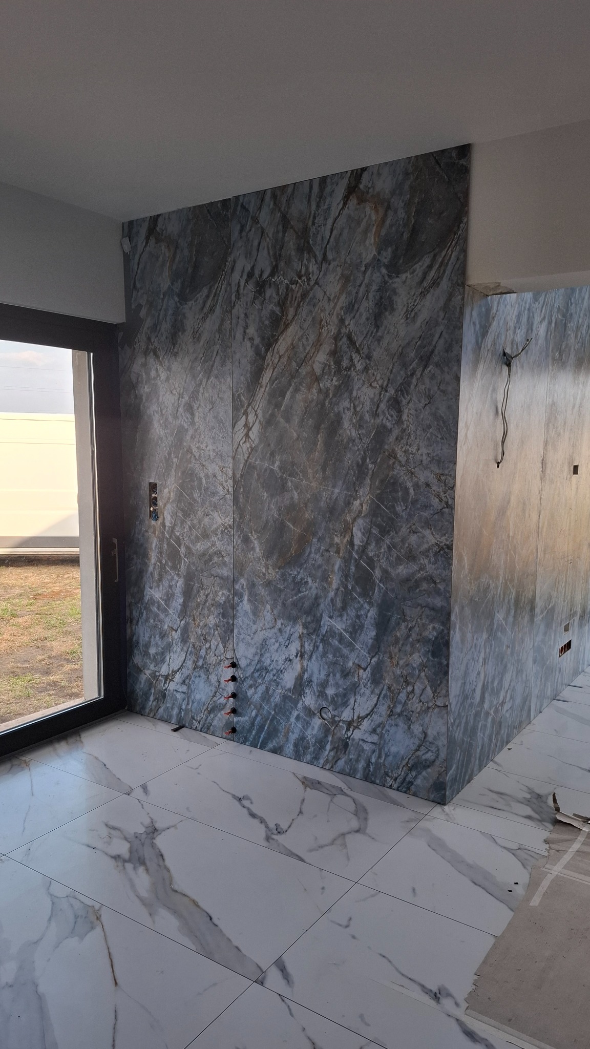 Ściana i podłoga wyłożone płytkami imitującymi marmur w Stepnicy. Widoczne fugi, okno z widokiem na zewnątrz oraz wystające kable elektryczne.