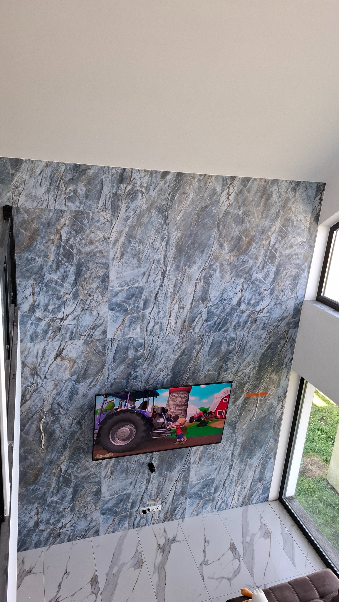 Ściana i podłoga wyłożone płytkami imitującymi marmur w odcieniach szarości i bieli. Na ścianie wisi telewizor. Widok z góry, częściowo widoczne okno i sofa.