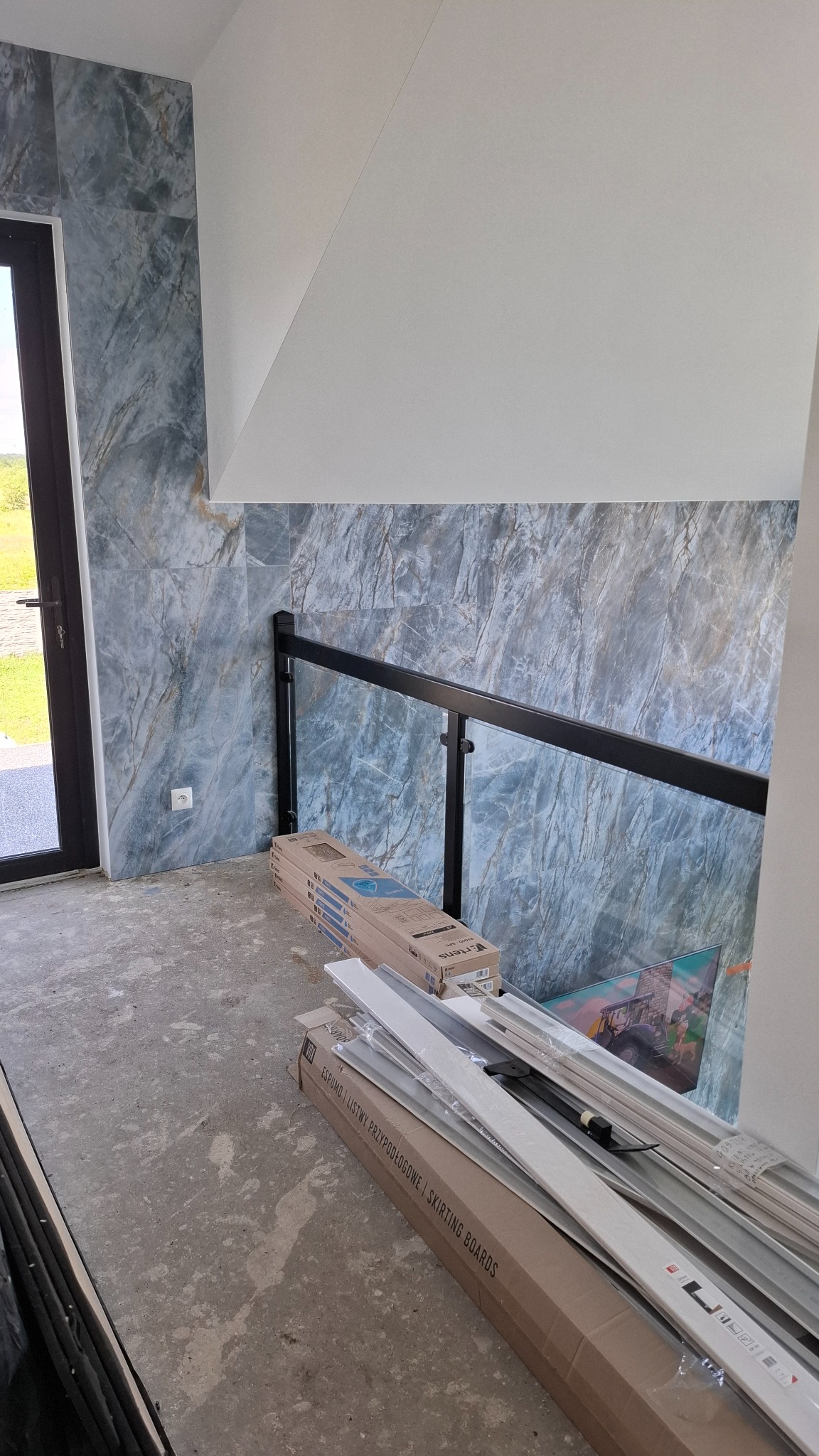 Ściana wykończona marmurowymi płytkami, czarna barierka ze szkłem, biała ściana pod skosem, kartony z listwami przypodłogowymi na surowej podłodze.