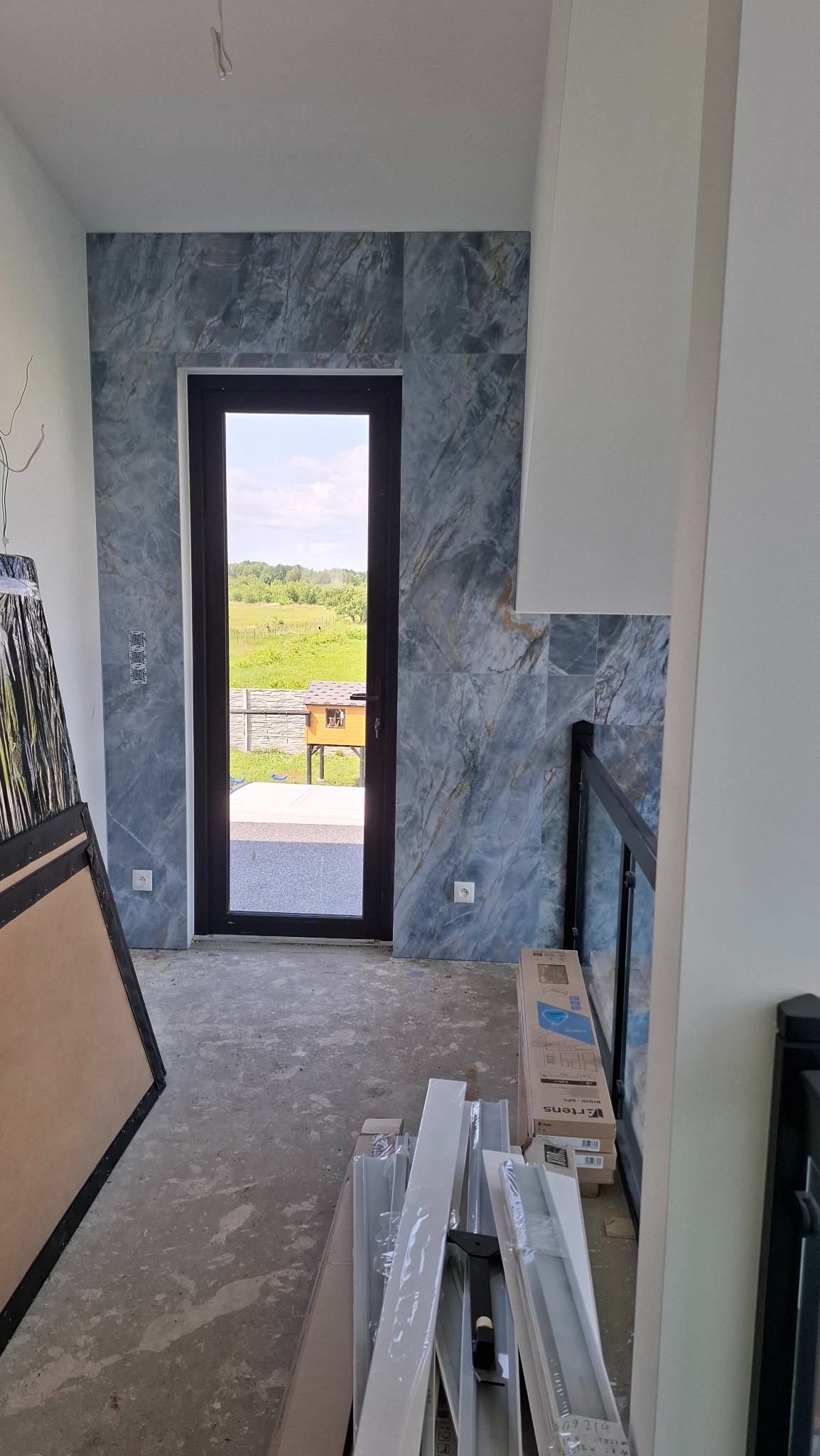 Ściana wyłożona marmurowymi płytkami w odcieniach szarości i błękitu wokół drzwi balkonowych. Widoczne materiały budowlane i surowa podłoga.