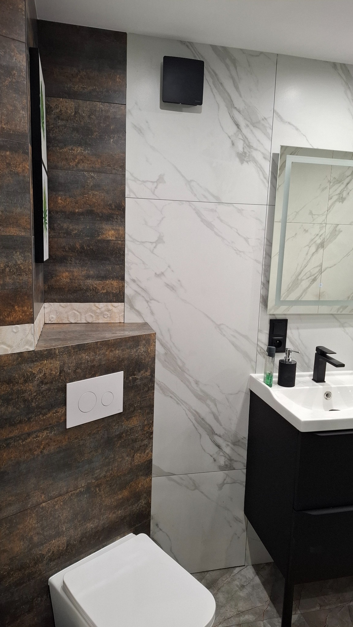 Nowoczesna łazienka z brązowo-białymi płytkami, czarną armaturą i podświetlanym lustrem. Widoczny fragment toalety i umywalka z szafką.