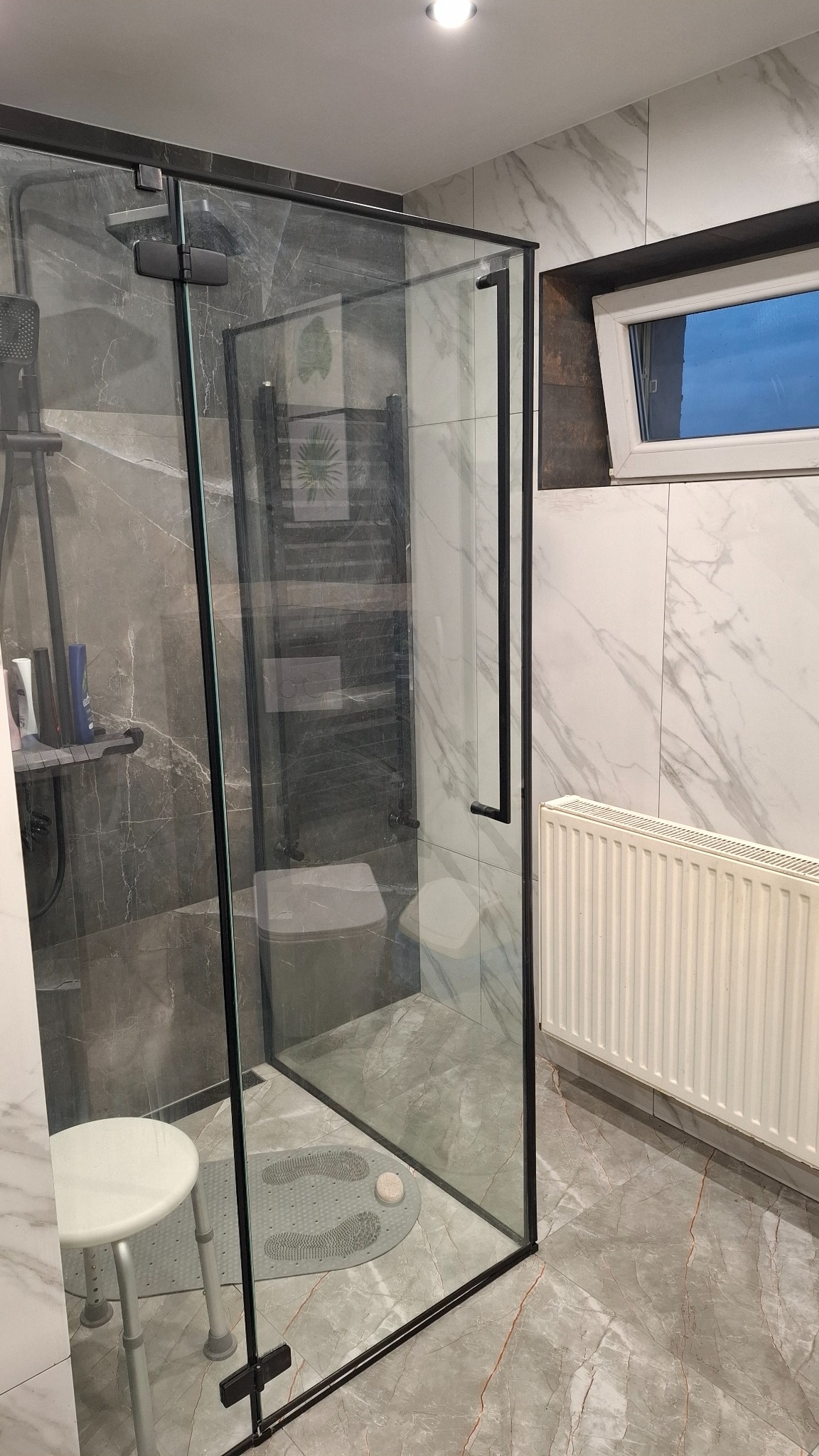 Nowoczesna kabina prysznicowa z czarnymi profilami i marmurowymi płytkami w łazience z grzejnikiem i oknem.