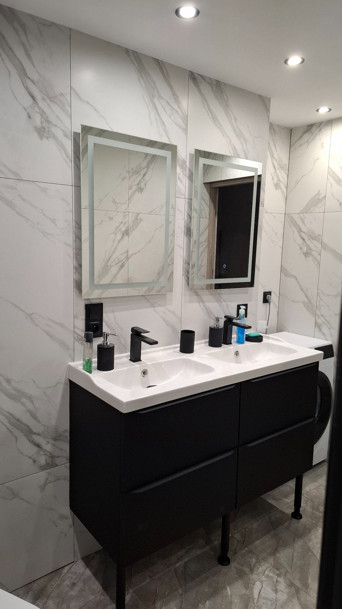 Nowoczesna łazienka z podwójną umywalką na czarnej szafce, marmurowe płytki na ścianach i podświetlane lustra. Widoczna pralka i akcesoria łazienkowe.