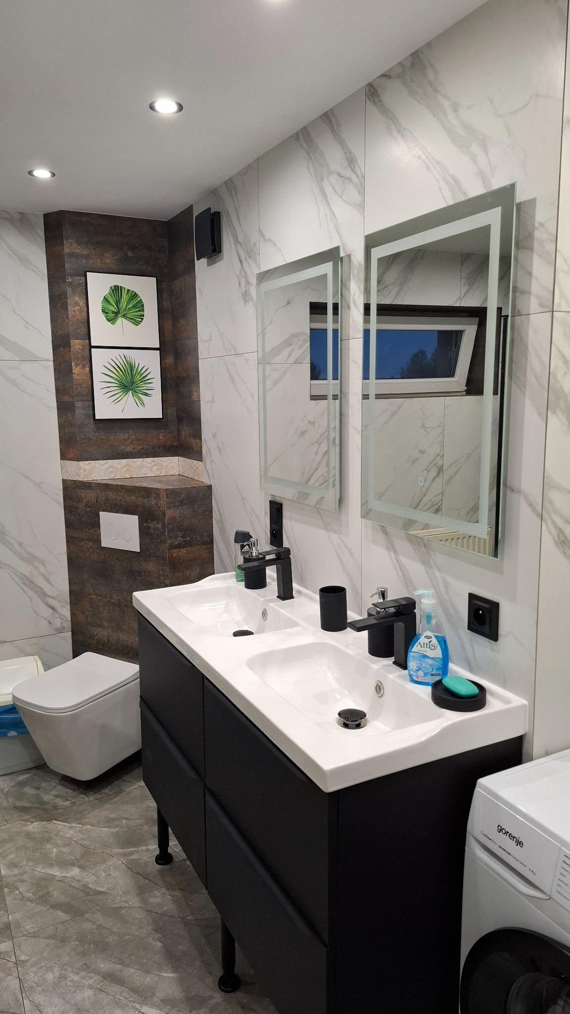 Nowoczesna łazienka z podwójną umywalką na czarnej szafce, marmurowe płytki, lustra z podświetleniem i akcenty w postaci obrazków z motywem liści.