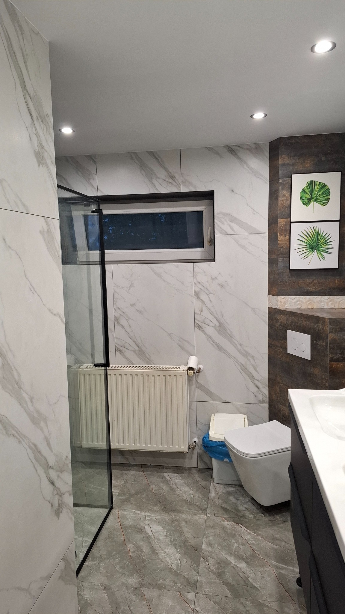 Nowoczesna łazienka z marmurowymi płytkami, kabiną prysznicową, grzejnikiem i wiszącą toaletą. Minimalistyczny design z akcentami zieleni w dekoracjach.