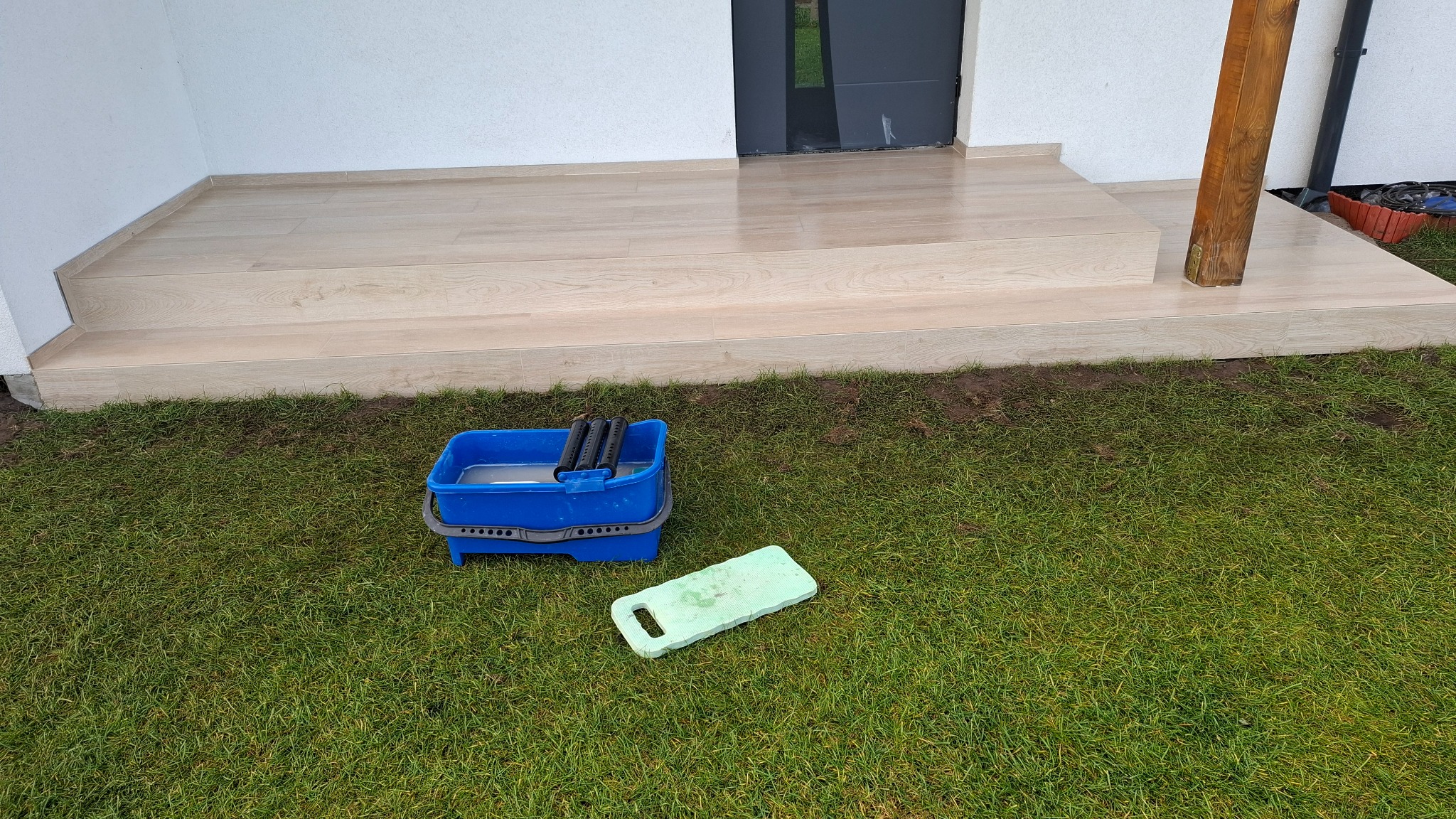Jasne płytki imitujące drewno na schodach wejściowych, z niebieskim wiadrem i podkładką na trawie. Widoczny fragment drewnianego słupa i drzwi.