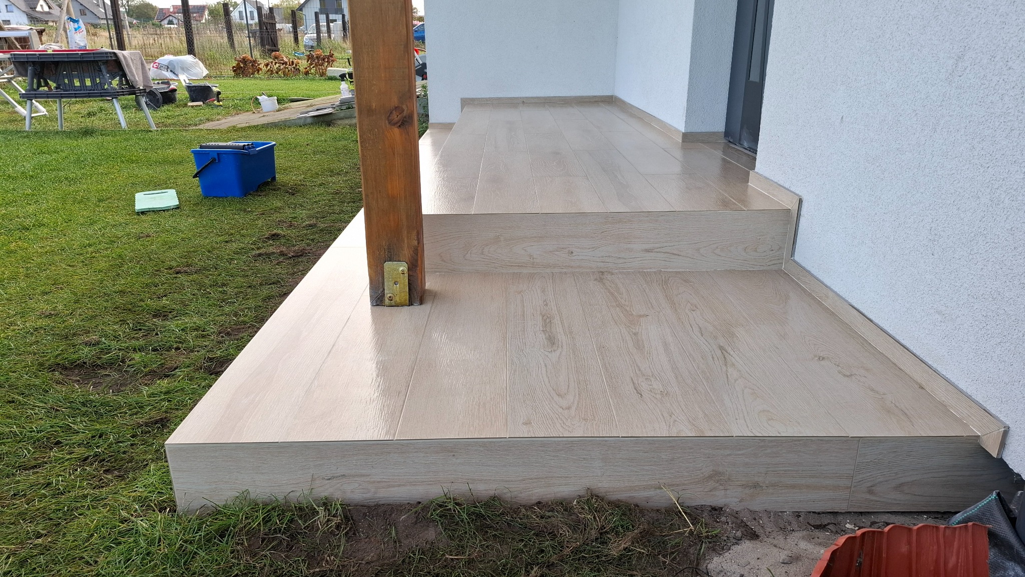 Wykończony taras z jasnych płytek imitujących drewno, z widocznym słupem podtrzymującym z żółtymi elementami mocującymi. Na trawie wiadro i gąbka.