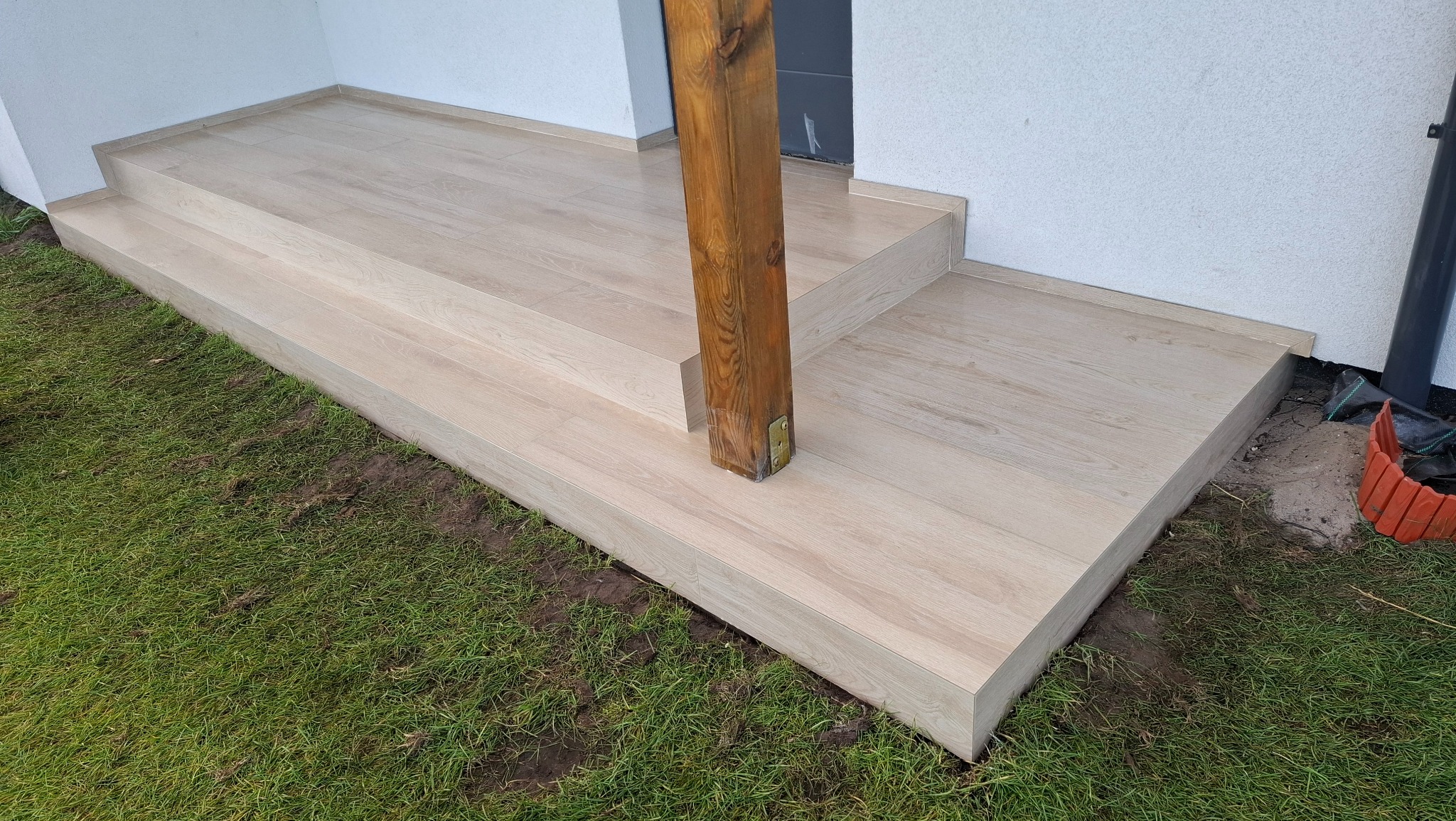 Wykończone płytkami schody wejściowe z drewnianym słupem, otoczone trawnikiem. Jasne płytki imitujące drewno, schludne wykończenie krawędzi. Widok z poziomu gruntu.