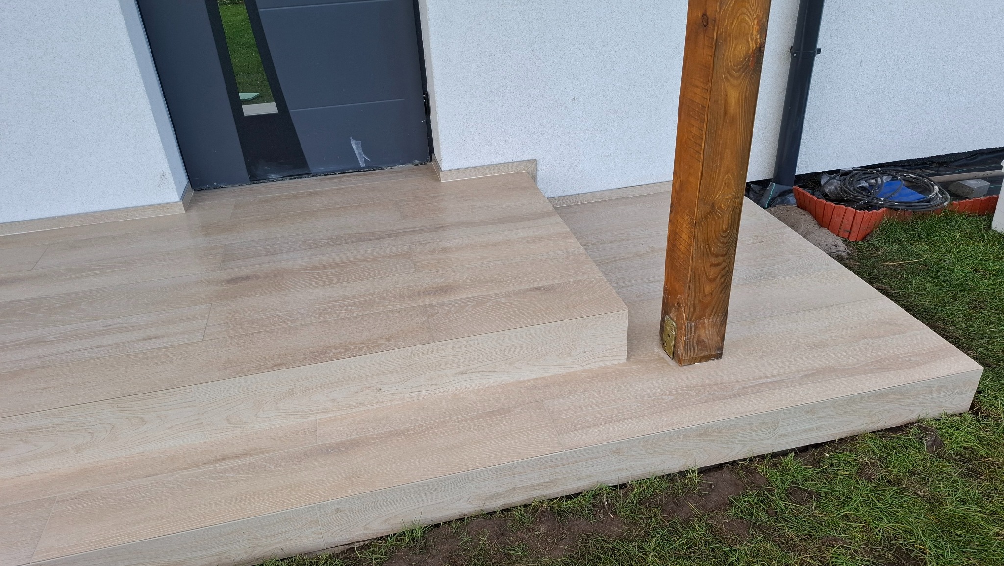 Wykończone płytkami schody wejściowe z drewnopodobnym wzorem, prowadzące do drzwi. Widoczny fragment drewnianego słupa i trawnika.