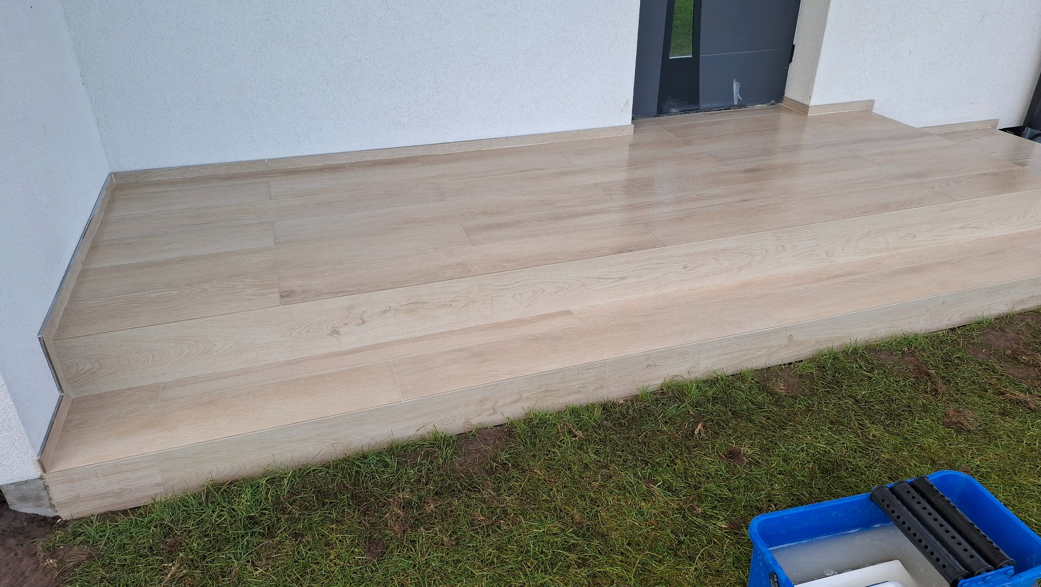 Jasne, drewnopodobne płytki na schodach wejściowych, z widocznym fragmentem trawnika i narzędziami glazurniczymi w rogu. Minimalistyczny design.
