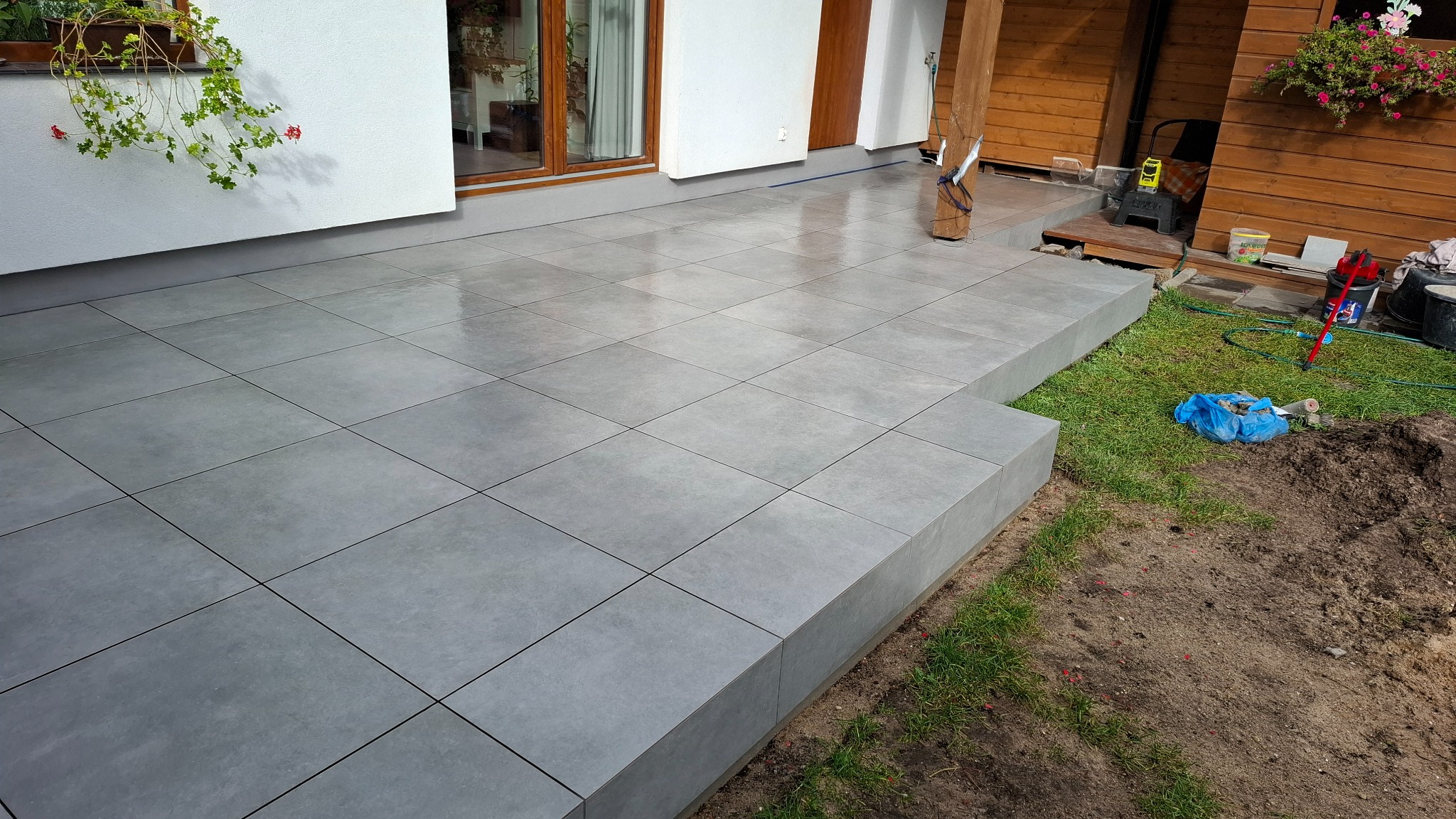 Szara, kwadratowa, błyszcząca podłoga z płytek na zewnątrz budynku, z widocznym fragmentem trawnika i elementami drewnianej elewacji. Widok z niskiej perspektywy.